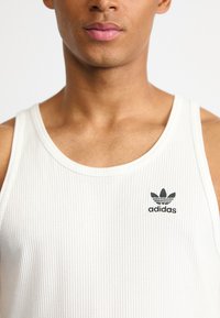 Bela rebrasta majica brez rokavov z okroglim izrezom. Na prsih je črn logo Adidas. Gladka tekstura, minimalističen dizajn.