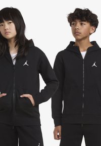 Μαύρες φούτερ ζακέτες με φερμουάρ, κατασκευασμένες από βαμβακερό μίγμα, με λευκό λογότυπο της Jordan, μπροστινές τσέπες και ραμμένα μανίκια. Μοντέλα που στέκονται προφίλ.