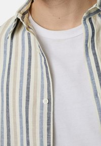 Camicia a righe con bottoni, caratterizzata da strisce orizzontali beige, azzurro chiaro e blu navy. Sotto si vede una maglietta bianca a collo tondo. Materiale in misto cotone.