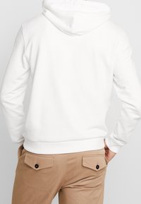 Vit hoodie med känguruficka, ribbstickade muddar och fåll. Kombinerad med beige byxor med platta bakfickor med knappdetaljer.