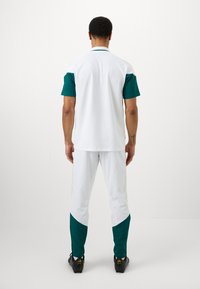 Άνδρας που στέκεται με την πλάτη στην κάμερα, φορώντας άσπρα και πράσινα αθλητικά ρούχα και μαύρα ποδοσφαιρικά παπούτσια, σε λευκό φόντο.