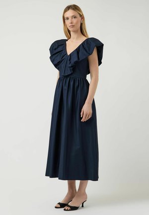Femme portant une robe midi bleu marine avec de grandes épaules à volants et un col en V, associée à des talons ouverts noirs, debout devant un fond uni.