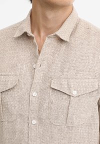 Camicia beige con bottoni, realizzata in una tessitura testurizzata, con due tasche frontali e bottoni bianchi. Design con colletto e vestibilità casual.