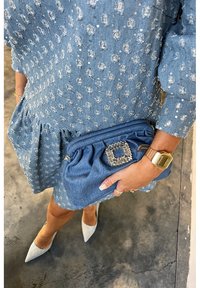 Femme en robe bleue texturée tenant une pochette bleue assortie avec une boucle en strass, portant une montre digitale dorée et des chaussures blanches pointues sur un sol en béton.