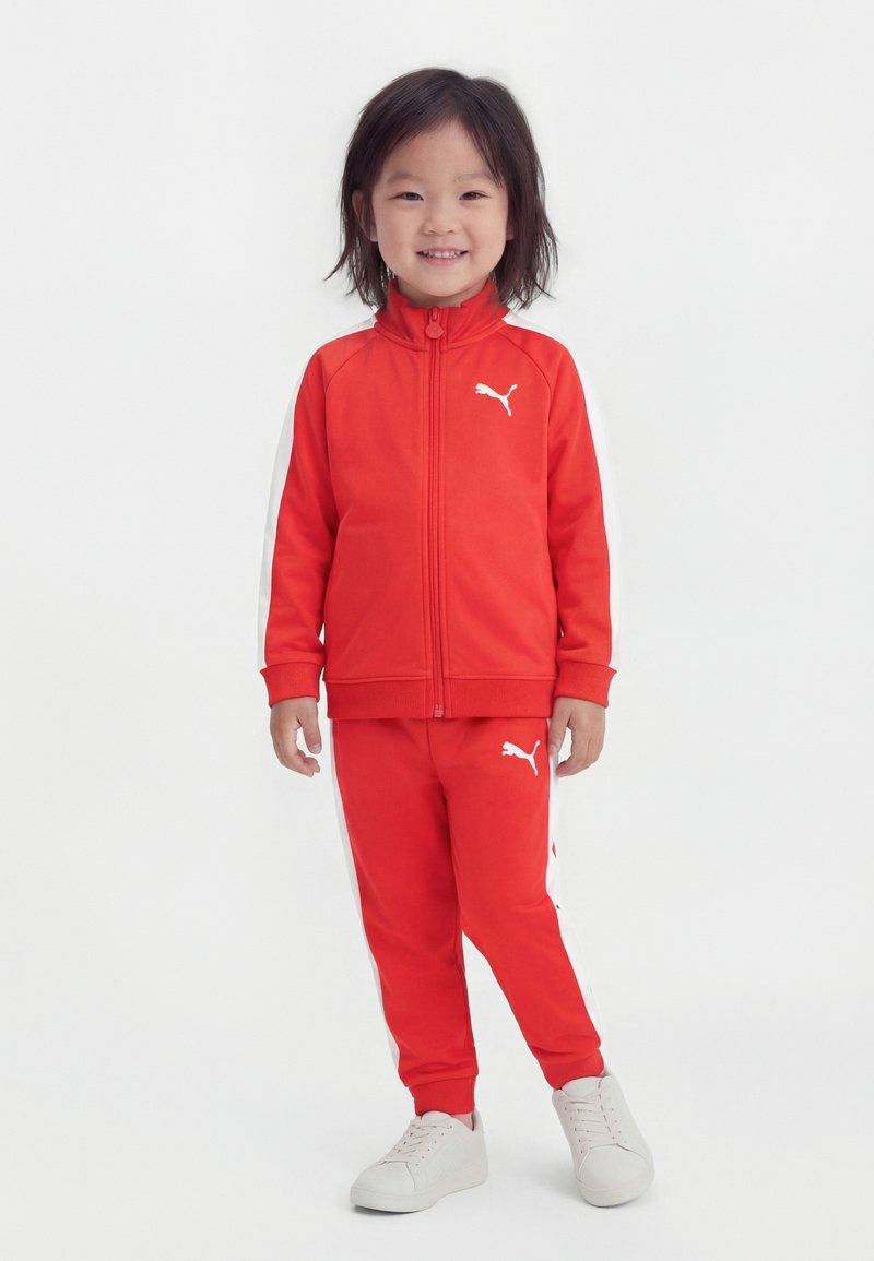 Kind lächelt, trägt einen roten Puma-Trainingsanzug mit weißem Logo und weißen Streifen, steht vor weißem Hintergrund mit weißen Turnschuhen.