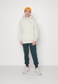 Hoodie grigio chiaro con tasca frontale e un piccolo logo, abbinato a pantaloni cargo verde scuro e sneakers bianche, e un berretto arancione brillante.