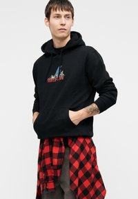 Crna dukserica s prednjim kangaroo džepom, koja sadrži šareni grafički logo. Nosena preko crveno-crne karirane flanel košulje.