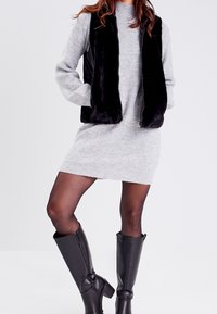 Veste en fausse fourrure noire portée sur une robe grise côtelée, associée à des bottes en cuir noires montant jusqu'aux genoux. Textures lisses et silhouette moderne.