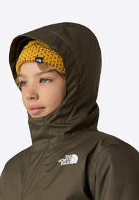 Vanntett jakke i olivengrønt med høy krage og justerbar hette; har en hvit logo på brystet, brukt sammen med en gul strikkelue.