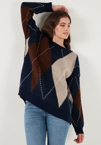Navy, brown, and beige argyle-patterned oversized sweater with a V-neck and ribbed cuffs, paired with light blue jeans.