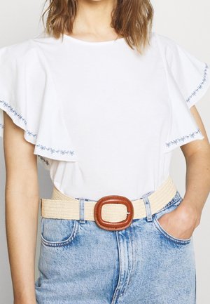 Femme portant un haut blanc à manches volantes avec une broderie florale bleue sur les manches, un jean en denim clair et une ceinture tissée beige avec une boucle marron.