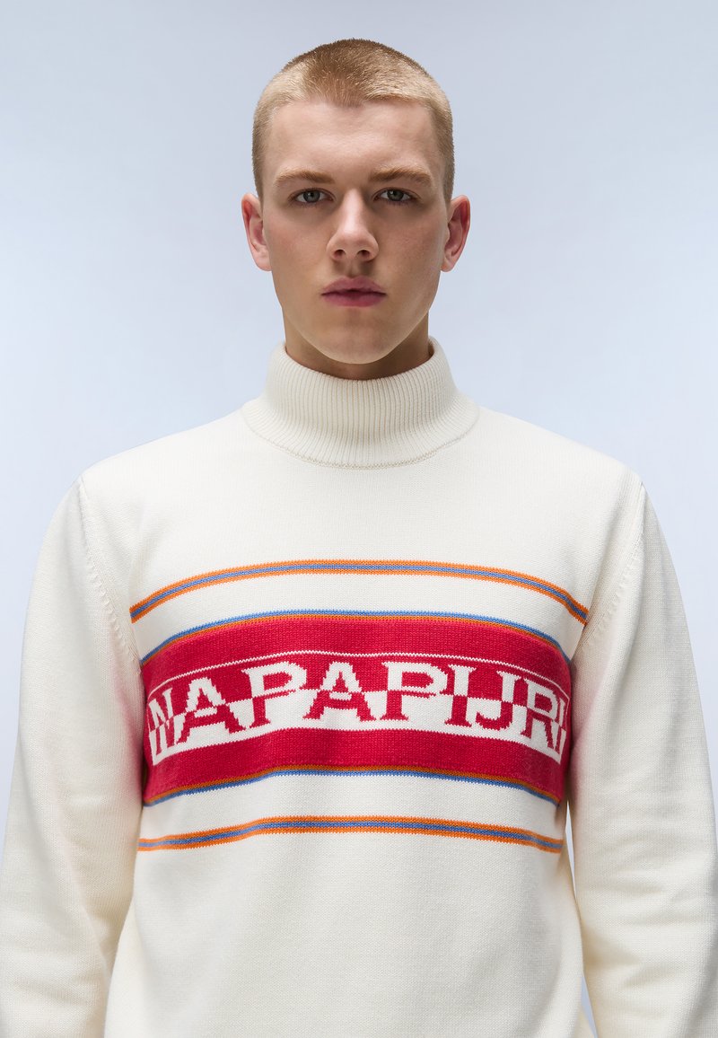 Biały sweter z wysokim kołnierzem, ozdobiony czerwoną poziomą taśmą z napisem "NAPAPIJRI" w bieli, wzbogacony kolorowymi paskami powyżej i poniżej.