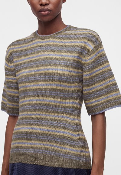 Femme portant un pull en maille à manches courtes avec des rayures horizontales dans des tons vert olive, violet et jaune.