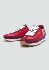 Un par de zapatillas rojas y burdeos con cordones blancos y una marca, suelas de goma beige y acentos azules, colocadas sobre un fondo gris claro.