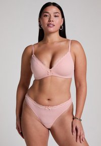 Lichtroze beha en bijpassende string, gemaakt van geribbelde stof. Kenmerkt kantafwerking met een klein strikje in het midden en verstelbare bandjes.