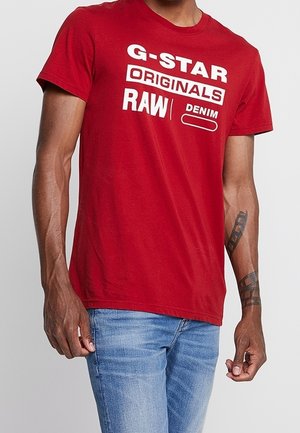 Czerwona bawełniana koszulka z odważnym białym napisem: "G-STAR ORIGINALS RAW DENIM". Okrągły dekolt, krótkie rękawy i luźny krój.