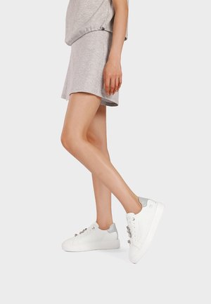 Donna con gonna e top grigi, in piedi su una gamba, che indossa sneakers bianche con tacco argento e lacci decorativi.