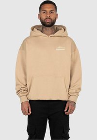Beige hoodie met een voorzak, geribbelde manchetten en een logo-detail op de borst. Zacht materiaal met een ontspannen pasvorm.