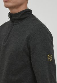 Sudadera de forro polar gris con cuello alto y media cremallera. Presenta un logotipo amarillo en la manga izquierda, textura suave y diseño ajustado.