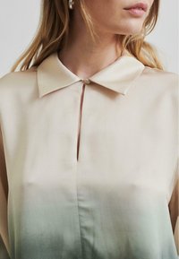 Blusa di seta di colore beige chiaro, con colletto classico, chiusura con bottoni davanti e un effetto ombre che sfuma in un verde tenue nella parte inferiore.