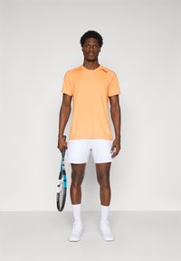Oranges Sport-T-Shirt mit kurzen Ärmeln, kombiniert mit weißen Shorts. Das Model hält einen Tennisschläger und trägt weiße Sneaker und Socken.