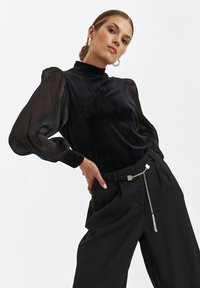 Czarna aksamitna bluzka z prześwitującymi puffkami, wysokim dekoldem i gładką teksturą. Połączona z czarnymi spodniami o wysokim stanie z dekoracyjnym łańcuchem.