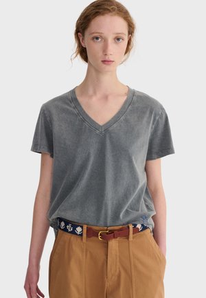 Junge Person mit hellem Teint und roten Haaren, die ein ausgewaschenes graues T-Shirt mit V-Ausschnitt trägt, das in beige Hosen gesteckt ist, dazu ein marineblauer Gürtel mit weißen Blattmustern.