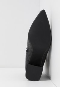 Chaussure noire à bout pointu avec semelle texturée et talon bloc placée debout contre un coin blanc.