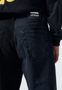 Pantalones vaqueros de mezclilla negra con corte recto, que cuentan con dos bolsillos traseros y una etiqueta rectangular que dice "DRDENIM" en la cintura.