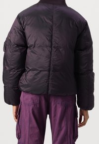 Fialová péřová bunda s prošívaným designem, žebrovaným límcem a bočními kapsami. Zadní strana má hladký povrch a minimální detaily.