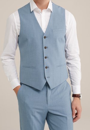 Homme portant un gilet de costume bleu clair et un pantalon assorti avec une chemise blanche et une montre-bracelet marron, debout avec une main dans la poche.