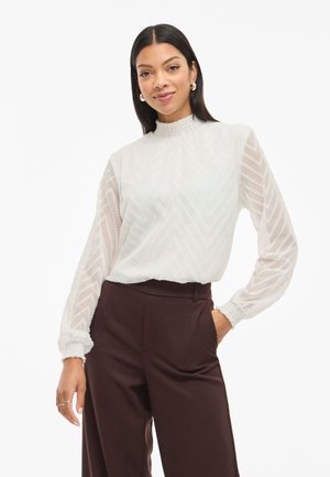 White blouse with textured chevron pattern, high ruffled neckline, and sheer long sleeves. Paired with dark brown tailored pants.