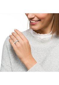 Sölvring med en texturerad yta, bärs på en hand. Personen har på sig en ljusgrå tröja med en volangkrage under.