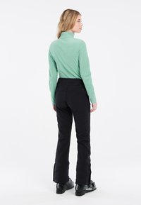 Top lungo a maniche con collo alto di colore verde menta, abbinato a pantaloni da sci neri con caviglie con zip e stivali da sci neri.