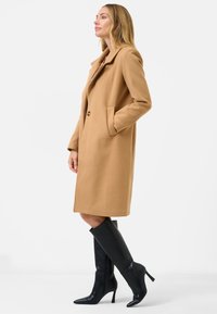 Beige wollen mix jas met een geknipt revers, enkele knoopsluiting en zijzakken. Draag het met zwarte knieketse laarzen met hak.