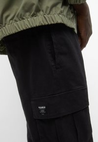 Zwarte cargobroek met een gestructureerde afwerking, voorzien van een zijzak en een labelpatch, gecombineerd met een olijfgroene elastische jaszoom.