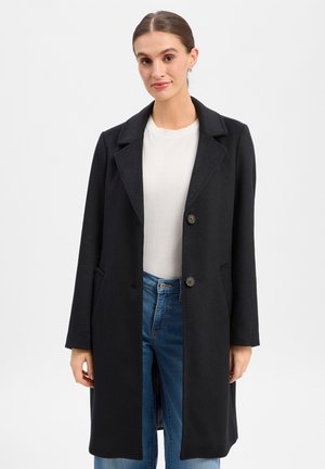 Woman wearing a black buttoned coat over a white top and blue jeans, standing against a plain light background.