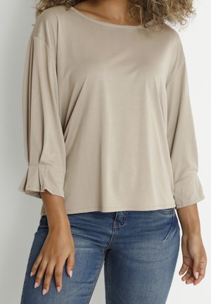 Femme portant un haut ample beige à manches longues avec des poignets évasés et un jean en denim bleu, debout les mains détendues sur les côtés.