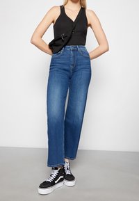 Jeans in denim blu a vita alta con gamba dritta, abbinati a un top nero senza maniche e sneakers nere alte. Include una borsa a tracolla nera.