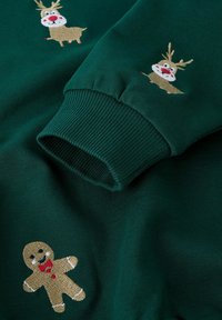 Ciemnozielona tkanina z haftowanym reniferem i piernikiem, z akcentem na prążkowane mankiety rękawów.
