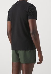 Camiseta negra de manga corta hecha de algodón, con cuello redondo y un detalle de logo en la parte posterior, combinada con pantalones cortos verdes.