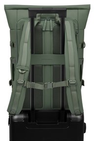 Rugzak met een groene stoffen buitenkant, gaasdetails, verstelbare schouderbanden en een uitschuifbare zwarte handgreep, ontworpen voor bevestiging aan een koffer.