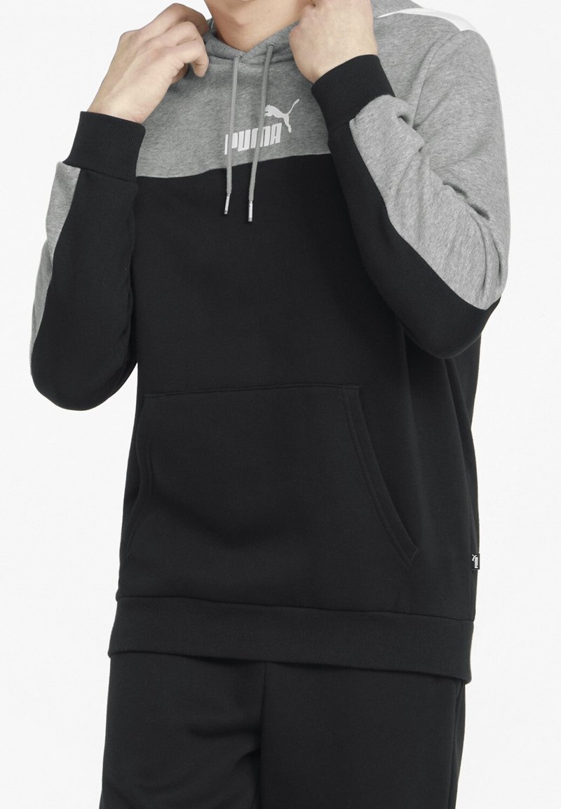 Personne ajustant la capuche d'un sweat à capuche Puma noir et gris avec poche avant et cordons.