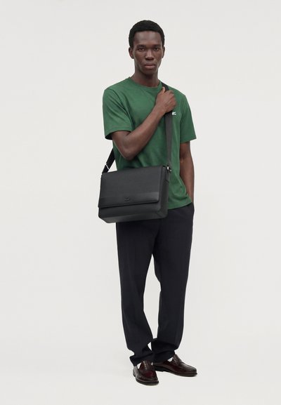 Un homme porte un sac besace noir en bandoulière par-dessus un T-shirt vert et un pantalon noir, sur un fond clair uni.
