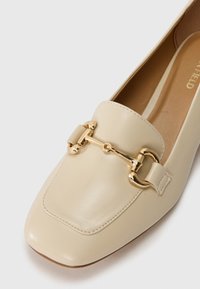 Beige nahkaa loafer, jossa on neliömäinen varvas, kultasävytetty yksityiskohta, sileä pinta ja brodeerattu koristelu vampissa. Litteä pohja.