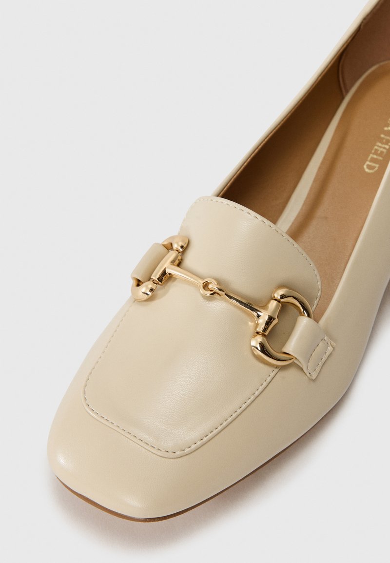 Mocassin en cuir beige avec bout carré, accent en métal doré, texture lisse et détail cousu sur le devant. Semelle plate.