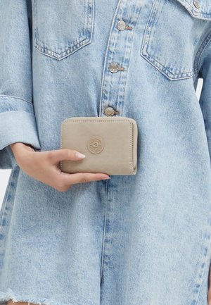 Portefeuille beige texturé avec un logo arrondi, tenu contre une chemise en denim clair. Fermeture zippée avec des bords lisses et une forme compacte.