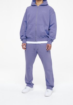 Sweat à capuche violet zippé avec poches avant, assorti à un pantalon de survêtement violet. T-shirt blanc visible en dessous, porté avec des baskets blanches.