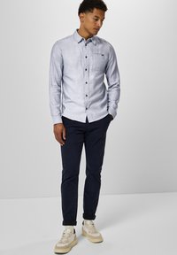 Camicia blu chiaro con bottoni e taschino sul petto, abbinata a pantaloni blu scuro e sneakers color crema. La camicia presenta un tessuto morbido e testurizzato.