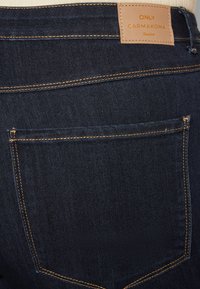 Jean em denim azul escuro com um rótulo de marca em couro, costura contrastante e um detalhe de um único bolso traseiro.